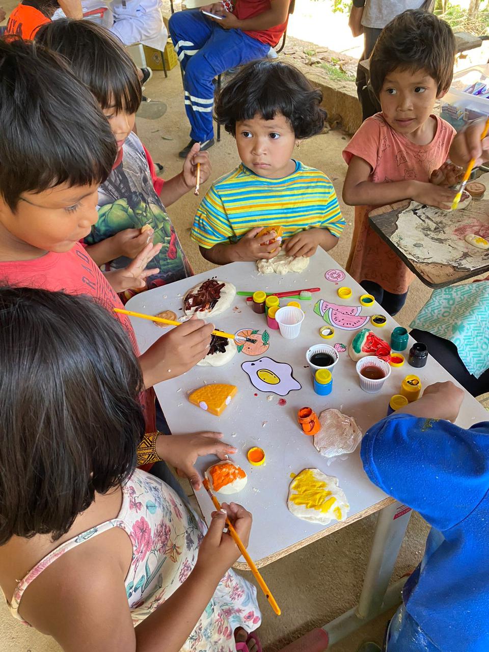 Na imagem, aparecem cinco crianças indígenas pintando massinhas de modelar que representam alimentos saudáveis e processados, em ação de orientação alimentar